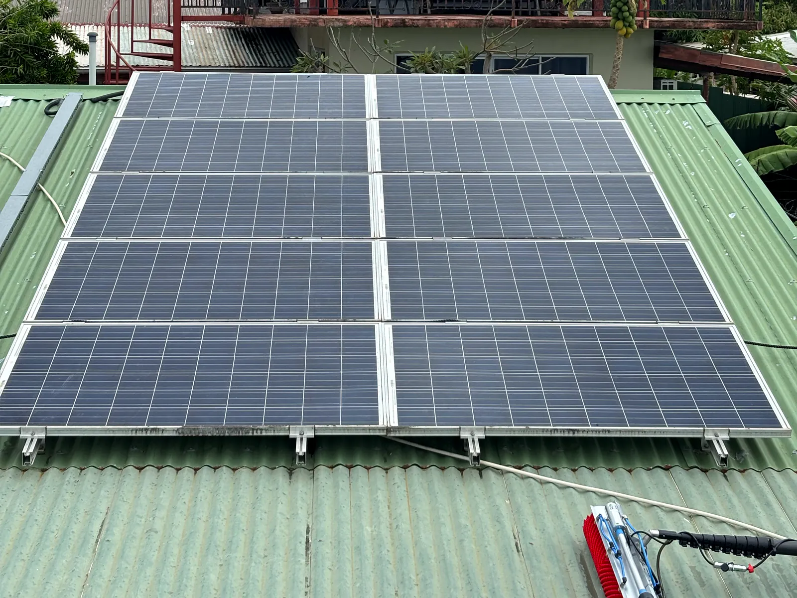 Rangées de panneaux photovoltaïques sur une toiture en tôle verte, perche et brosse au premier plan, maison au fond