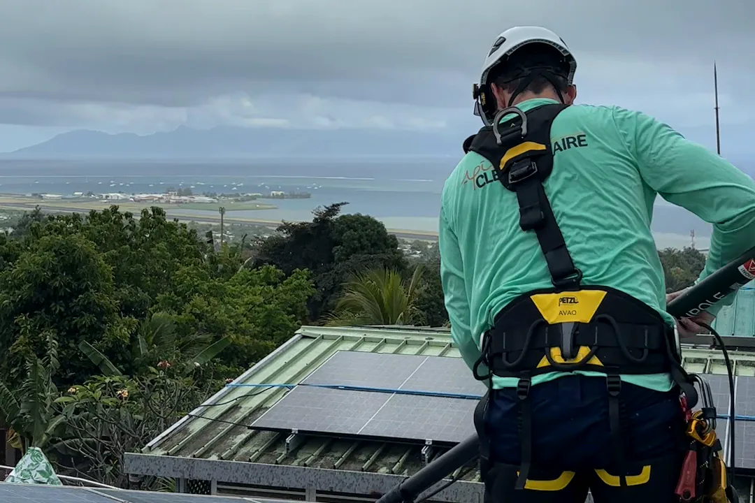 Technicien de dos en harnais nettoyant des panneaux solaires à la brosse rotative sur un toit à Tahiti, lagon au fond