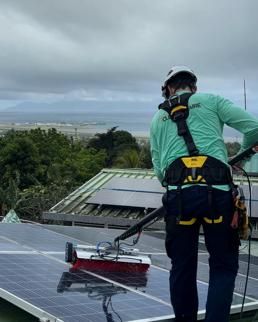 Technicien de dos en harnais nettoyant des panneaux solaires à la brosse rotative sur un toit à Tahiti, lagon au fond