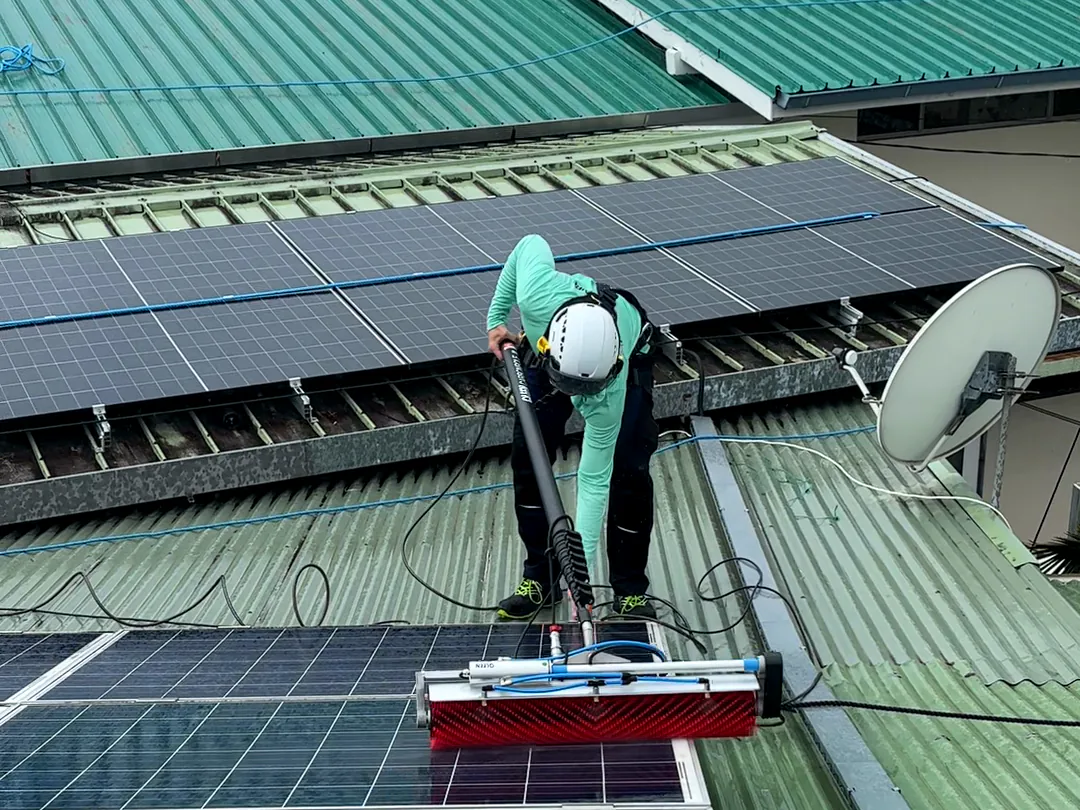 Technicien casqué penché sur sa perche, debout sur une toiture en tôle verte entre deux rangées de panneaux solaires